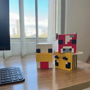 Pode incluir: Tr&ecirc;s figuras de personagens do Minecraft, um frango, uma vaca e uma abelha, feitas de pequenos blocos quadrados. O frango &eacute; branco com um bico amarelo e uma crista vermelha. A vaca &eacute; vermelha com manchas brancas e olhos pretos. A abelha &eacute; amarela com listras pretas e olhos pretos.
