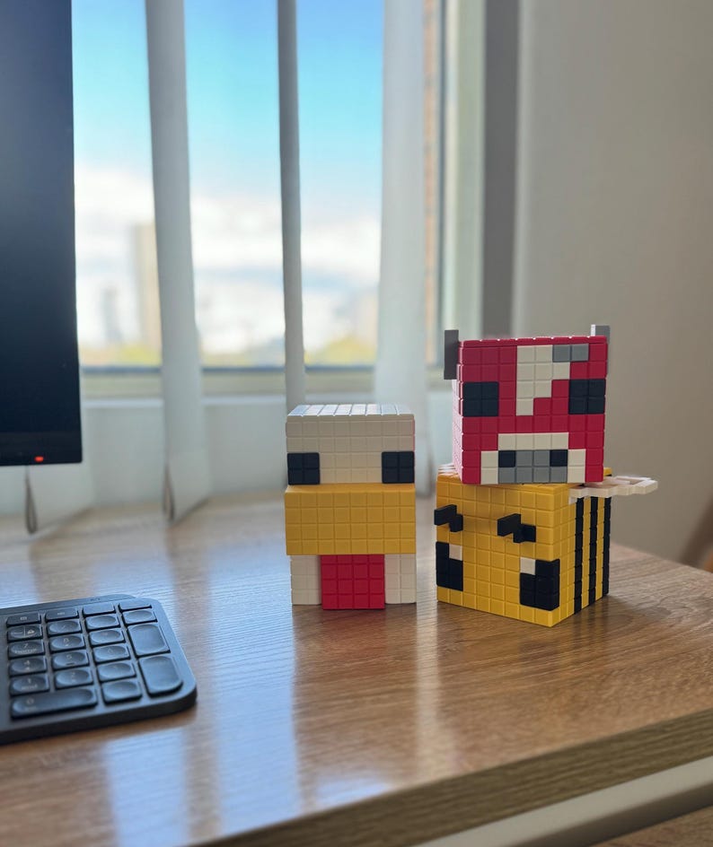 Pode incluir: Tr&ecirc;s figuras de personagens do Minecraft, um frango, uma vaca e uma abelha, feitas de pequenos blocos quadrados. O frango &eacute; branco com um bico amarelo e uma crista vermelha. A vaca &eacute; vermelha com manchas brancas e olhos pretos. A abelha &eacute; amarela com listras pretas e olhos pretos.