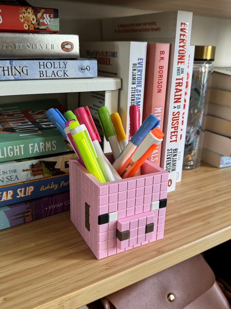 May include: A pink pig-shaped pencil holder made of plastic blocks. The holder has black eyes and a brown snout. It is filled with colourful pens and highlighters.