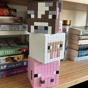 May include: A stack of three Minecraft-themed block toys. The top block is brown and white with a cow face, the middle block is white with a sheep face, and the bottom block is pink with a pig face.