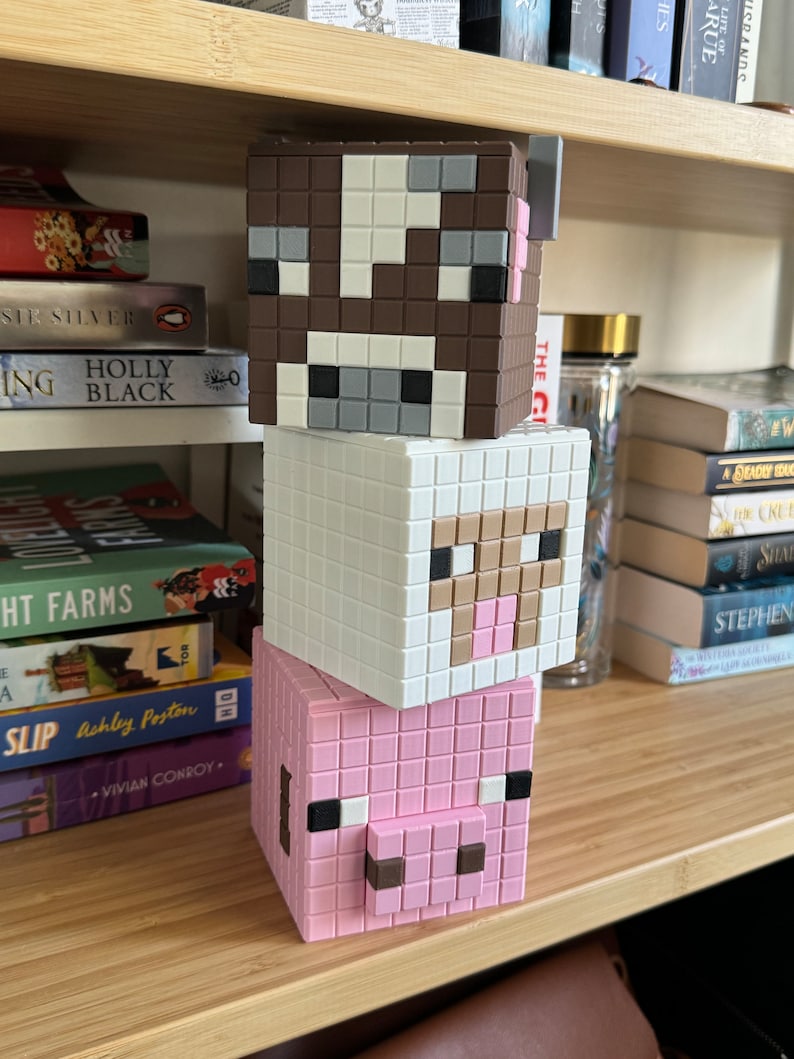 May include: A stack of three Minecraft-themed block toys. The top block is brown and white with a cow face, the middle block is white with a sheep face, and the bottom block is pink with a pig face.