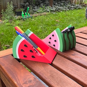 May include: Watermelon-shaped desk tidies in red, pink, green, and black. The organisers are made of a hard material and are shaped like watermelon slices. Pens and markers are inside the tidies. The tidies are on a wooden table.
