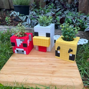Pode incluir: Tr&ecirc;s vasos de plantas com tema Minecraft, cada um com um personagem diferente: uma vaca vermelha e branca, um frango branco com bico amarelo e uma abelha amarela e preta. Os vasos s&atilde;o feitos de um material branco e t&ecirc;m um design pixelado.