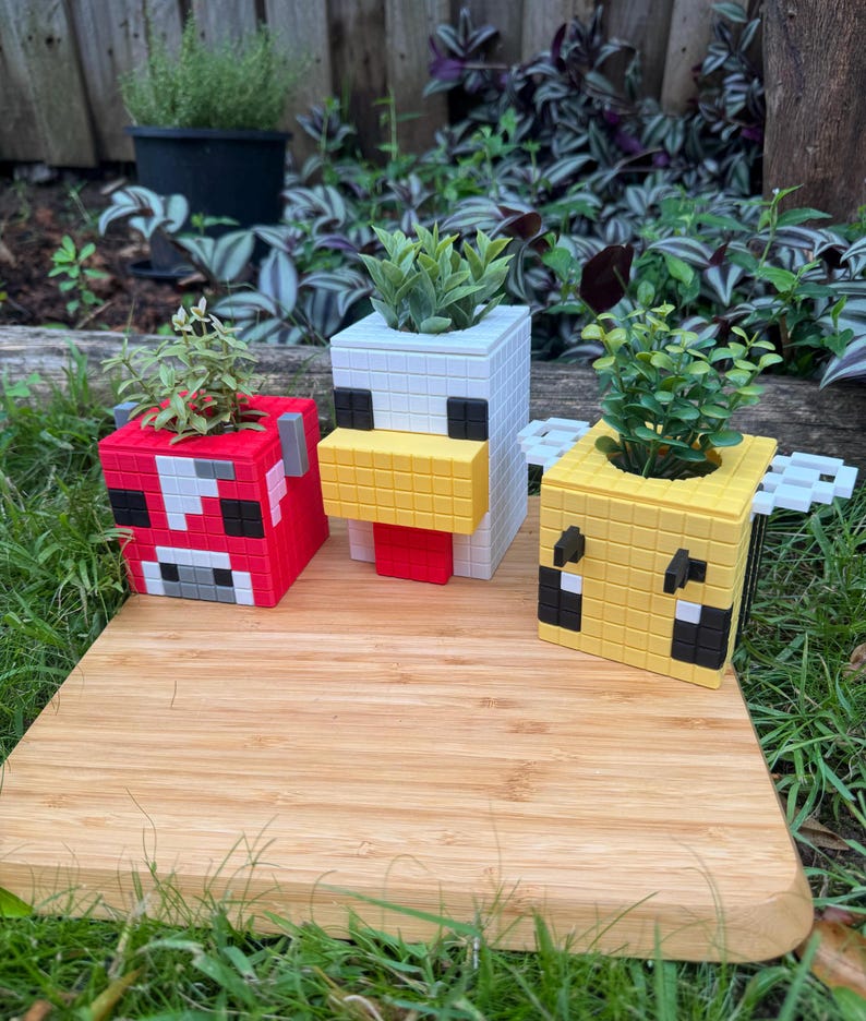 Pode incluir: Tr&ecirc;s vasos de plantas com tema Minecraft, cada um com um personagem diferente: uma vaca vermelha e branca, um frango branco com bico amarelo e uma abelha amarela e preta. Os vasos s&atilde;o feitos de um material branco e t&ecirc;m um design pixelado.