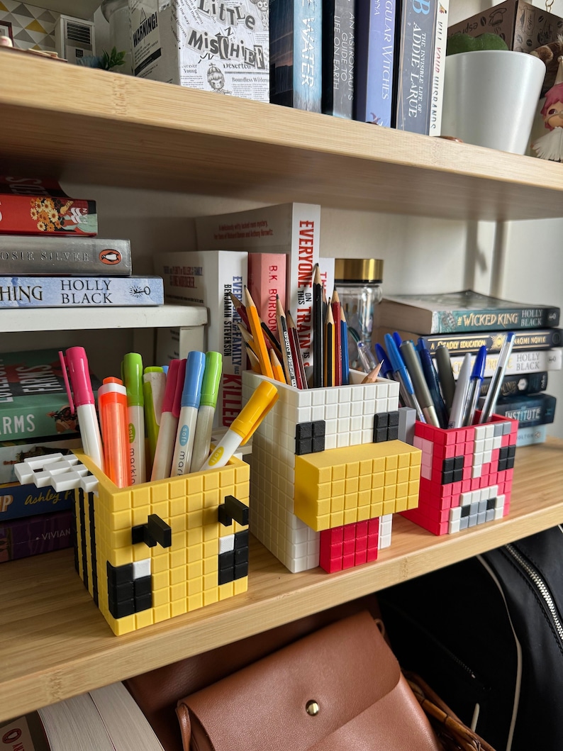 Pode incluir: Tr&ecirc;s porta-l&aacute;pis coloridos em forma de abelha, frango e porco. Os porta-l&aacute;pis s&atilde;o feitos de pl&aacute;stico e t&ecirc;m um design pixelado. Os porta-l&aacute;pis est&atilde;o cheios de canetas e l&aacute;pis.