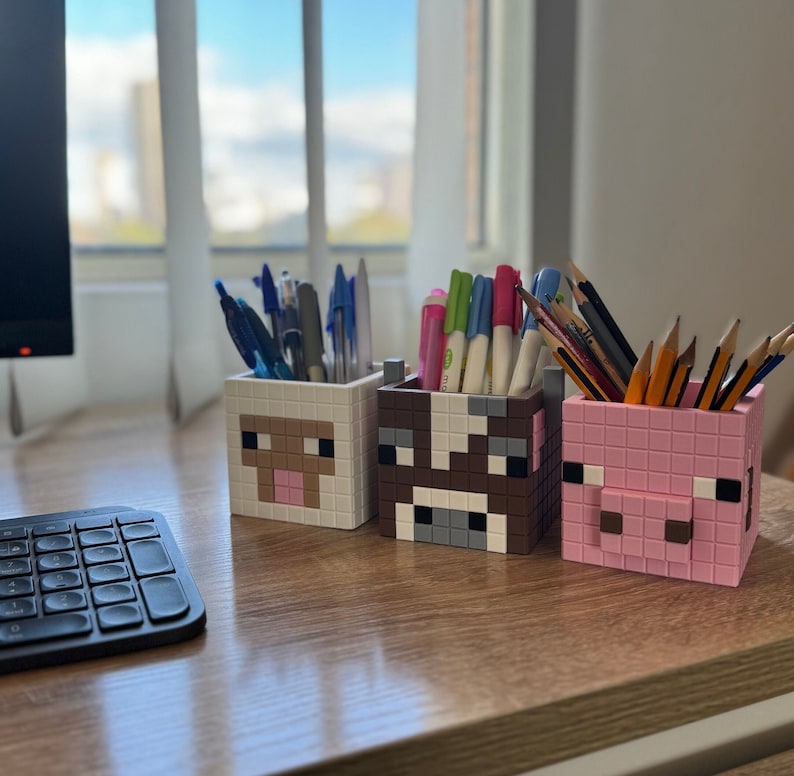 May include: Three colourful pencil holders shaped like Minecraft characters. The holders are made of plastic and feature a pig, a cow, and a sheep. The holders are filled with various coloured pens and pencils.