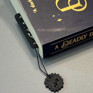 Może przedstawiać: Czarny zakładka z ozdobnym wzorem i czarną frędzlą zwisa z książki zatytułowanej "A Deadly Education" autorstwa Kiran Millwood Hargrave. Okładka książki jest ciemnoniebieska z złotymi literami.