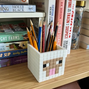 May include: A white pencil holder shaped like a Minecraft sheep with brown and pink details. The holder is filled with pencils of various colours.