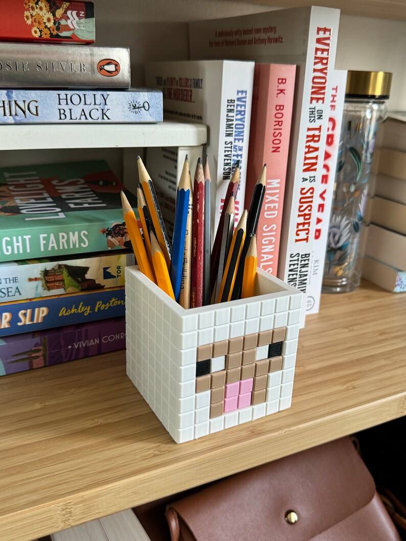 May include: A white pencil holder shaped like a Minecraft sheep with brown and pink details. The holder is filled with pencils of various colours.