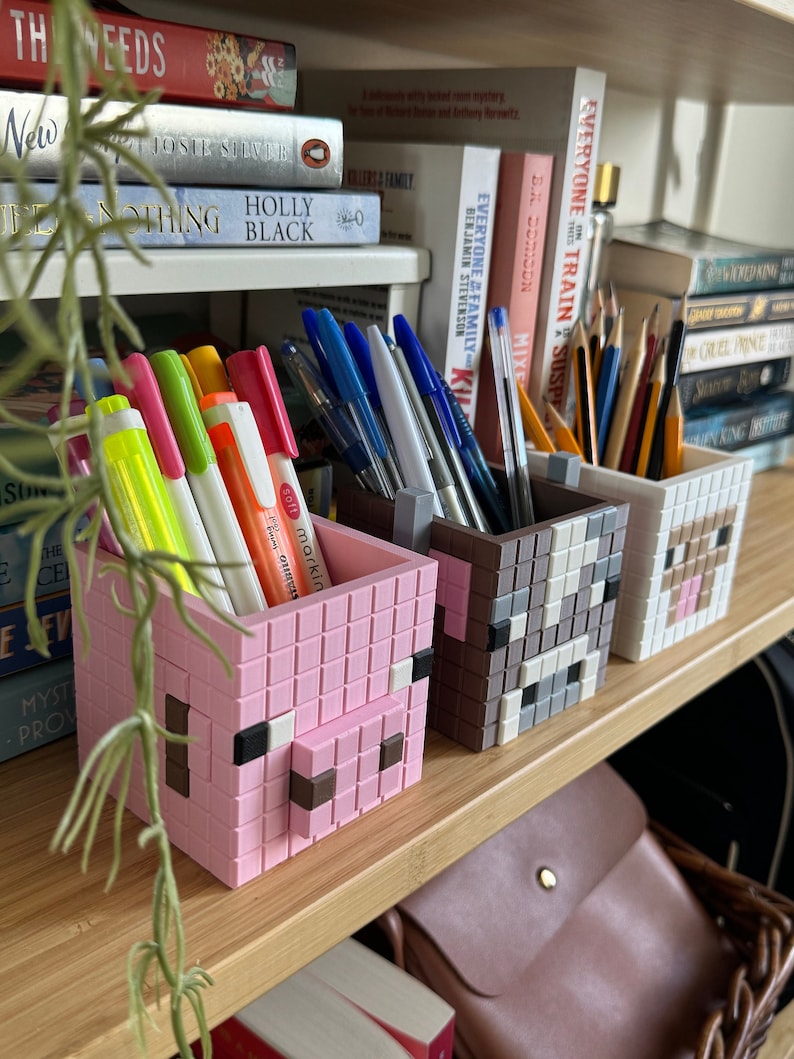 May include: Three Minecraft-themed pencil holders with pixelated animal faces, in pink, grey and brown. The holders are on a wooden shelf with books and a brown leather bag.