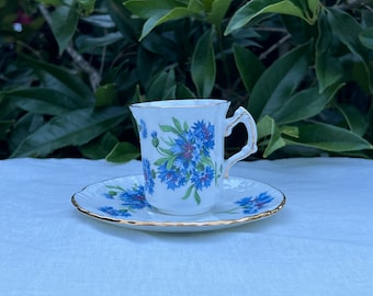 May include: A white teacup and saucer with a floral pattern of blue cornflowers. The cup has a gold rim and a handle. The saucer also has a gold rim.