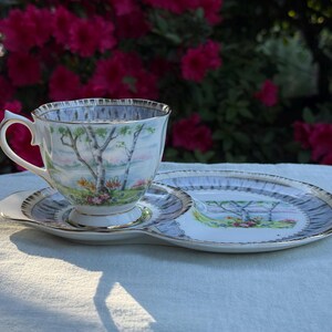 May include: A vintage teacup, saucer, and side plate set. The cup and plate feature a landscape scene with trees and flowers, with gold trim. The set is on a white surface, with a backdrop of pink flowers.