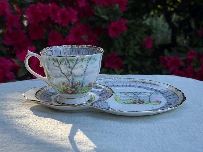 May include: A vintage teacup, saucer, and side plate set. The cup and plate feature a landscape scene with trees and flowers, with gold trim. The set is on a white surface, with a backdrop of pink flowers.