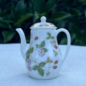 Puede incluir: Una tetera de porcelana blanca con un diseño floral con flores rosas y fresas rojas. La tetera tiene un borde dorado alrededor de la tapa y el pico.