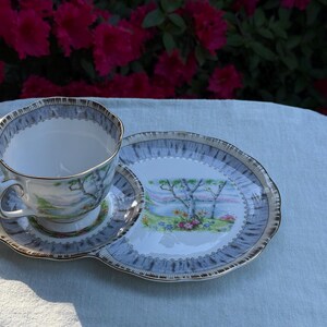 May include: A vintage teacup, saucer, and serving plate set. The teacup and saucer have a light blue and gold trim. The serving plate features a landscape scene with trees and flowers. The set is placed on a light blue tablecloth.