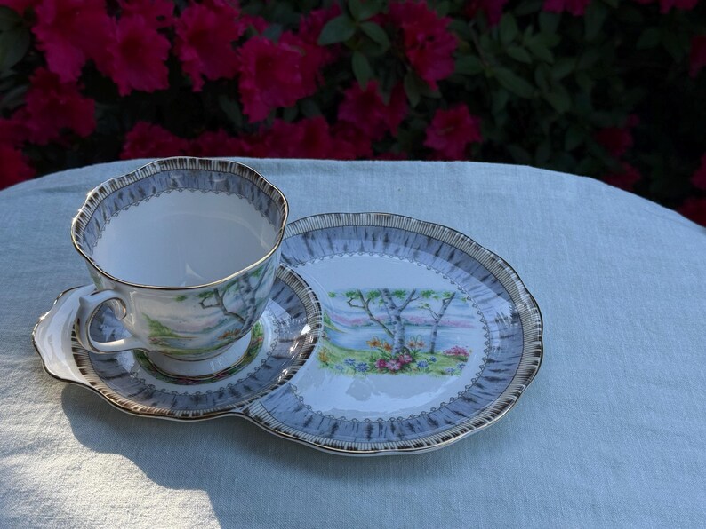 May include: A vintage teacup, saucer, and serving plate set. The teacup and saucer have a light blue and gold trim. The serving plate features a landscape scene with trees and flowers. The set is placed on a light blue tablecloth.