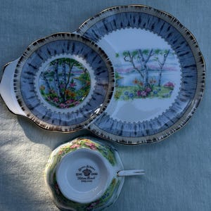 May include: A vintage tea set comprising a cup and two plates. The plates and cup have a blue and gold rim with a landscape scene. The cup is floral patterned. The set is made of bone china.