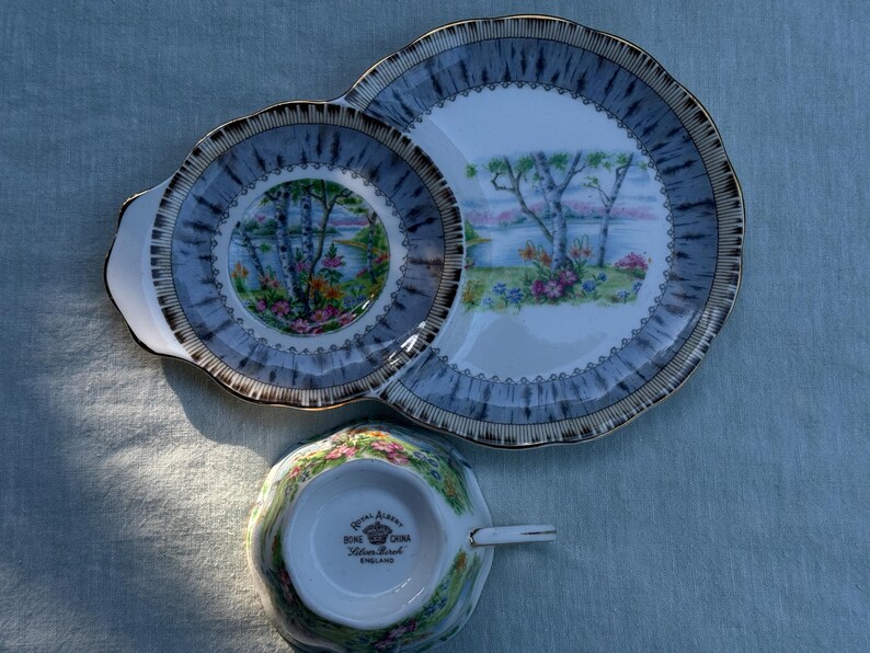 May include: A vintage tea set comprising a cup and two plates. The plates and cup have a blue and gold rim with a landscape scene. The cup is floral patterned. The set is made of bone china.