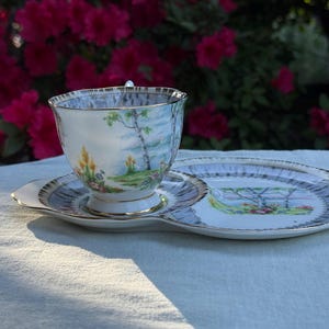 May include: A vintage teacup and saucer set featuring a landscape scene. The white porcelain cup and saucer have a gold trim. The saucer has a unique shape with a section for a biscuit or small treat.