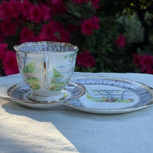 May include: A vintage teacup and saucer set with a landscape design. The cup and saucer have a white base with a gold trim and a light blue border. The landscape features trees, flowers, and a body of water. The set is placed on a white surface.