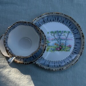 May include: A vintage teacup, saucer, and snack plate set. The set has a white interior with a blue and gold rim. The plate features a landscape scene with trees and flowers. The set is on a blue surface.