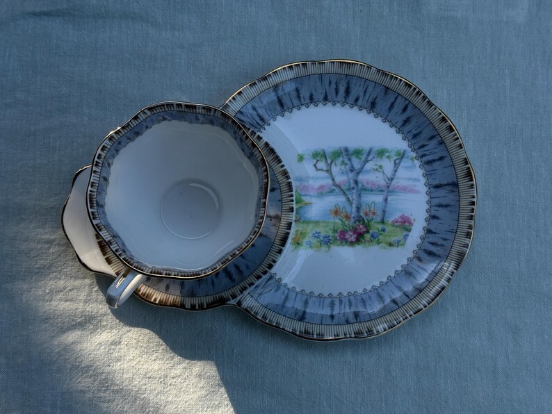 May include: A vintage teacup, saucer, and snack plate set. The set has a white interior with a blue and gold rim. The plate features a landscape scene with trees and flowers. The set is on a blue surface.