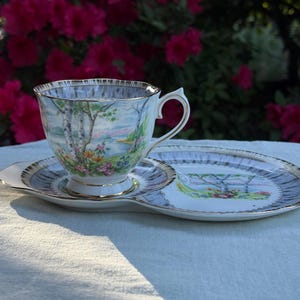 May include: A vintage teacup and saucer set with a landscape design featuring trees, flowers, and a body of water. The set has a gold trim and a matching plate. The background is blurred with pink flowers.