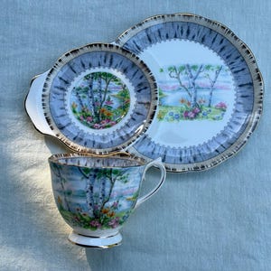 May include: A vintage tea set comprising a teacup, a saucer, and a serving plate. The set has a blue and gold rim with a landscape scene of trees, water, and flowers. The teacup and saucer have a handle.