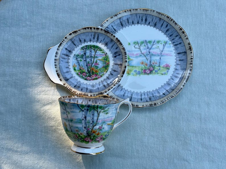 May include: A vintage tea set comprising a teacup, a saucer, and a serving plate. The set has a blue and gold rim with a landscape scene of trees, water, and flowers. The teacup and saucer have a handle.