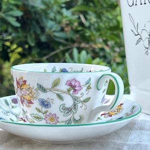 May include: A white teacup and saucer with a floral pattern in shades of pink, yellow, green, and blue. The cup has a green rim and a handle. The saucer has a scalloped edge.