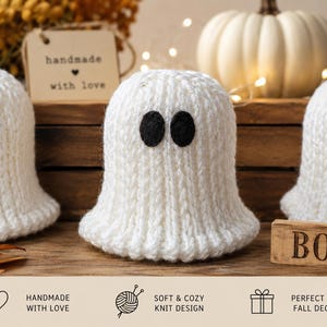 May include: Three white knitted ghost figures with black felt circular eyes. Each ghost has a ribbed texture and a rounded top. A small wooden tag reads handmade with love and a rectangular wooden block features the text BOO in black letters.