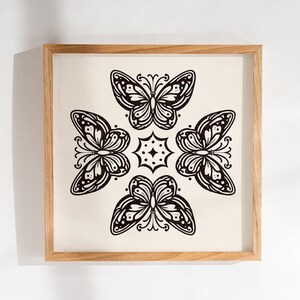 Puede incluir: Una impresión enmarcada con un diseño en blanco y negro de cuatro mariposas estilizadas dispuestas en un patrón simétrico. Las mariposas tienen detalles intrincados y se colocan sobre un fondo crema.