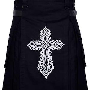 May include: Black kilt with a white, ornate cross design on the front. The kilt features pleats, belt loops, and leather straps. A gothic or alternative fashion item.