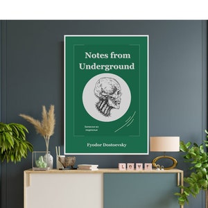 May include: A framed poster with the title "Notes from Underground" by Fyodor Dostoevsky. The poster features a green background with a skull illustration. The poster is displayed on a modern cabinet with plants and decorative items.