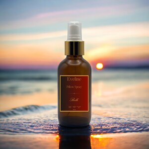 May include: A brown glass bottle with a gold lid and a white spray nozzle. The bottle is labeled "Eveline Pillow Spray Belle" with a red background and gold lettering. The bottle is sitting on a beach with a sunset in the background.