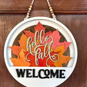 Peut inclure: Panneau rond en bois blanc avec l'inscription "Hello Fall WELCOME" en noir et beige. Le panneau présente des feuilles d'automne colorées dans des tons orange, rouge et jaune. Conçu pour être accroché à une porte ou un mur.
