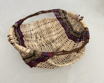 May include: Handwoven basket with a natural beige weave and a curved handle. The basket features accents of burgundy, blue, and olive green woven into the rim. The handle is made of a dark brown, twig-like material.