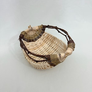 Puede incluir: Una cesta tejida a mano con un tejido natural de color claro. El borde y el asa están adornados con ramitas marrones oscuras y detalles tejidos. La cesta tiene forma redondeada y está sobre un fondo blanco.