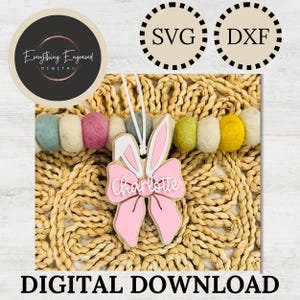 Può includere: Un ornamento a forma di coniglietto pasquale rosa e bianco con il nome "Charlotte", appeso su uno sfondo decorativo. L'immagine include anche il testo "DOWNLOAD DIGITALE" e i loghi dei file SVG e DXF.