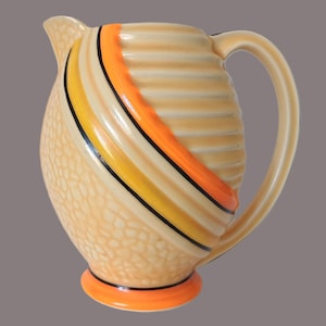 May include: A ceramic jug featuring a cream-coloured body with a textured pebble pattern on the lower section and horizontal ridges on the upper section. It is decorated with diagonal stripes in orange, yellow, and black, and has an orange base.