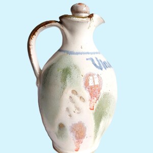 May include: A cream-coloured ceramic cruet with a handle and a lid. The surface features hand-painted green and orange abstract splotches, a blue horizontal line, and the partial blue text "Vin" written in a cursive script on the front side.