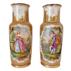 May include: A pair of antique porcelain vases with a gold rim and a painted scene of a man and woman in a garden. The vases have a cream-coloured top with gold accents and a tan base.