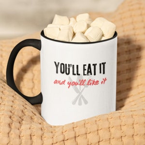 Può includere: Tazza in ceramica bianca con manico nero. La tazza ha il testo "You'll eat it and you'll like it" con un disegno di posate incrociate.