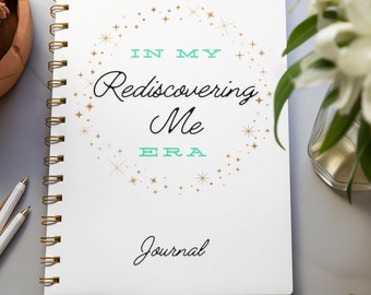 May include: A white journal with a gold spiral binding. The cover features a gold starburst design with the text "In My Rediscovering Me Era Journal".