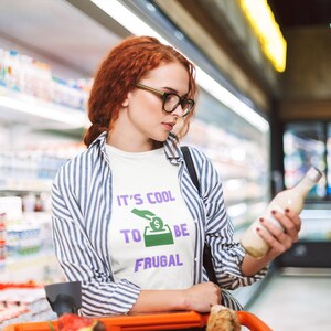 May include: A woman in a grocery store aisle is looking at a bottle of milk. She is wearing a white t-shirt with the text "It's cool to be frugal" printed on it. The text is in purple and green.