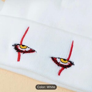 May include: White beanie with red and yellow embroidered eyes. The eyes are stylized and resemble a clown's eyes. The beanie is made of a soft, knit fabric.