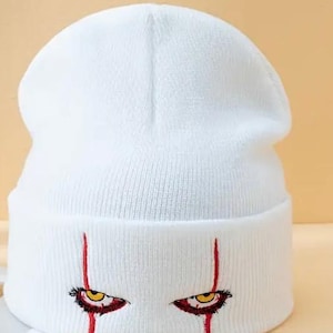 May include: White beanie with red and yellow embroidered eyes. The eyes have a menacing look. "Color: White" is written below the beanie.