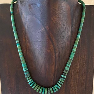 Puede incluir: Un collar de cuentas turquesa con cuentas de tamaño graduado. El collar se muestra en un soporte de madera.