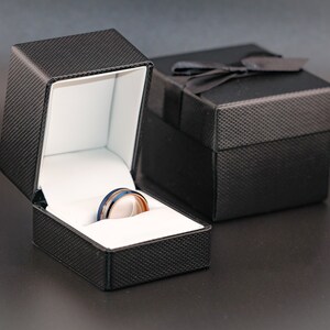 Puede incluir: Una caja de anillo negra con un interior blanco, abierta para revelar un anillo plateado y azul con una banda negra.