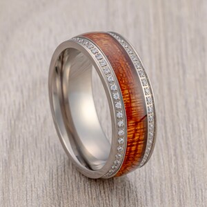 Puede incluir: Una banda de boda de plata con una incrustación de madera marrón y una fila de diamantes transparentes alrededor del borde.